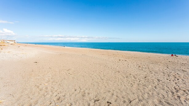 Een strand met een heldere lucht, een rustige zee en enkele mensen in het zand.