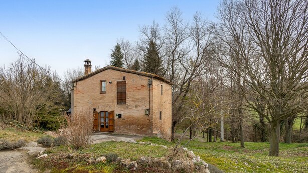 Une maison de campagne en briques, dotée d’une porte en bois, entourée d’arbres et bordée d’un chemin de pierres.