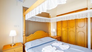 Ein ordentlich bezogenes Bett mit blauer Steppdecke, Holz-Kopfstück und Baldachin mit Spitze. Zwei Nachttische mit Lampen, ein Holznachtschränkchen und ein Kleiderschrank mit Spiegel.