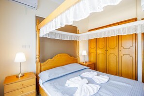 Ein ordentlich bezogenes Bett mit blauer Steppdecke, Holz-Kopfstück und Baldachin mit Spitze. Zwei Nachttische mit Lampen, ein Holznachtschränkchen und ein Kleiderschrank mit Spiegel.