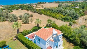 A two-story house with a red-tiled roof, surrounded by greenery and a view of the sea.