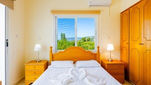 A bedroom with a bed, two bedside tables, a wardrobe, a window with a view of greenery, and an air conditioning unit.