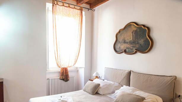 Ein ordentlich bezogenes Bett mit Kissen und gefalteten Handtüchern, ein Fenster mit durchsichtigen Vorhängen und ein an der Wand hängendes Bild.