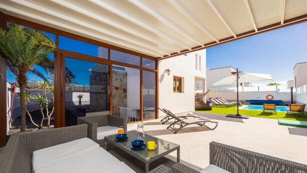 A covered outdoor area with wicker furniture, a glass table, and a pool.