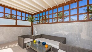 A covered outdoor seating area with wicker furniture and a glass table, large windows with a view of palm trees, and a tiled floor.