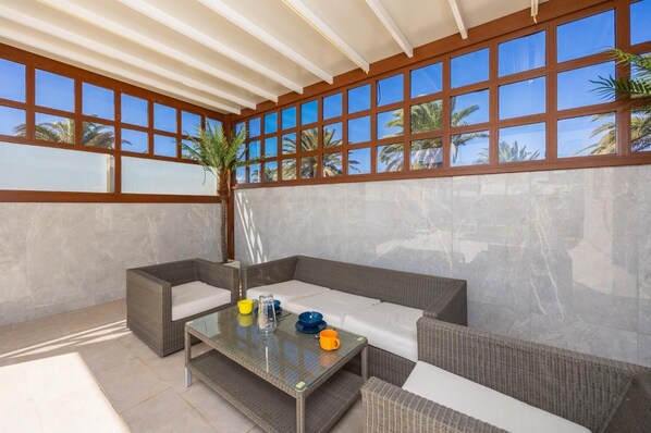 A covered outdoor seating area with wicker furniture and a glass table, large windows with a view of palm trees, and a tiled floor. - Las Caletas - Four Bedroom Villa, Sleeps 8 (Teguise)
