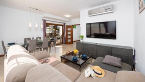 A modern living room with a sofa, coffee table, and TV. There is a dining area with a table and chairs, and a sliding glass door leading to a balcony.