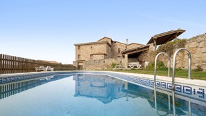 A swimming pool with clear blue water, surrounded by a wooden fence, and a stone building with a roofed patio area in the background.