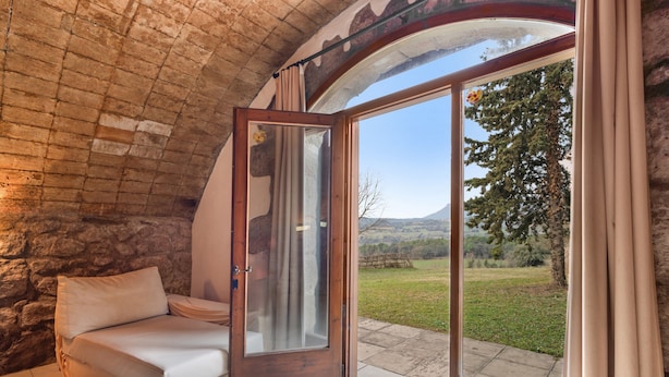 A room with a stone ceiling, a bed with a white mattress and pillow, a glass door leading outside, and a view of a grassy landscape.