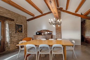 A dining area with a wooden table and white chairs, a chandelier, stone walls, and exposed wooden beams.