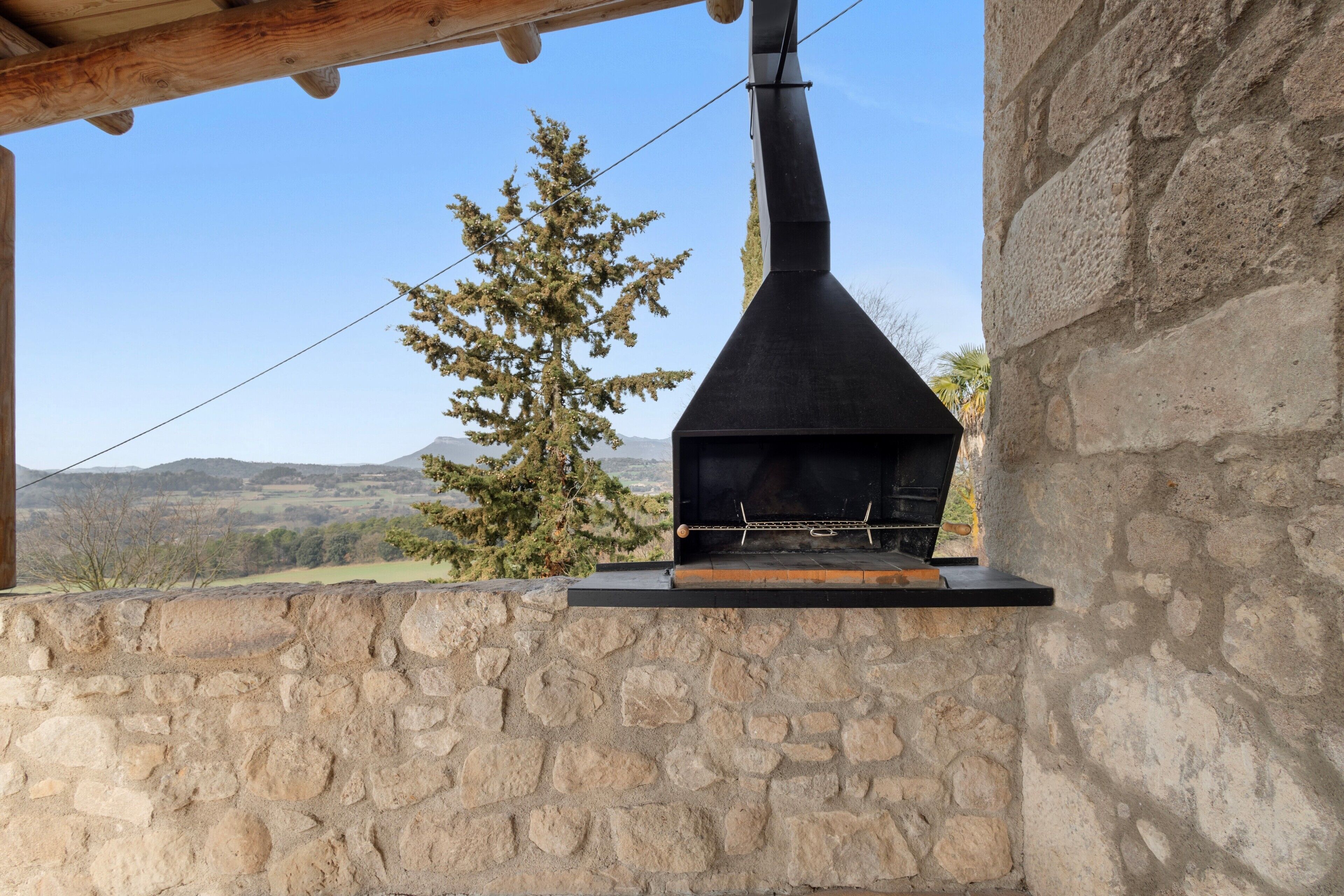 A stone wall with a wood-framed structure above it, a chimney, and a view of a tree and distant hills.