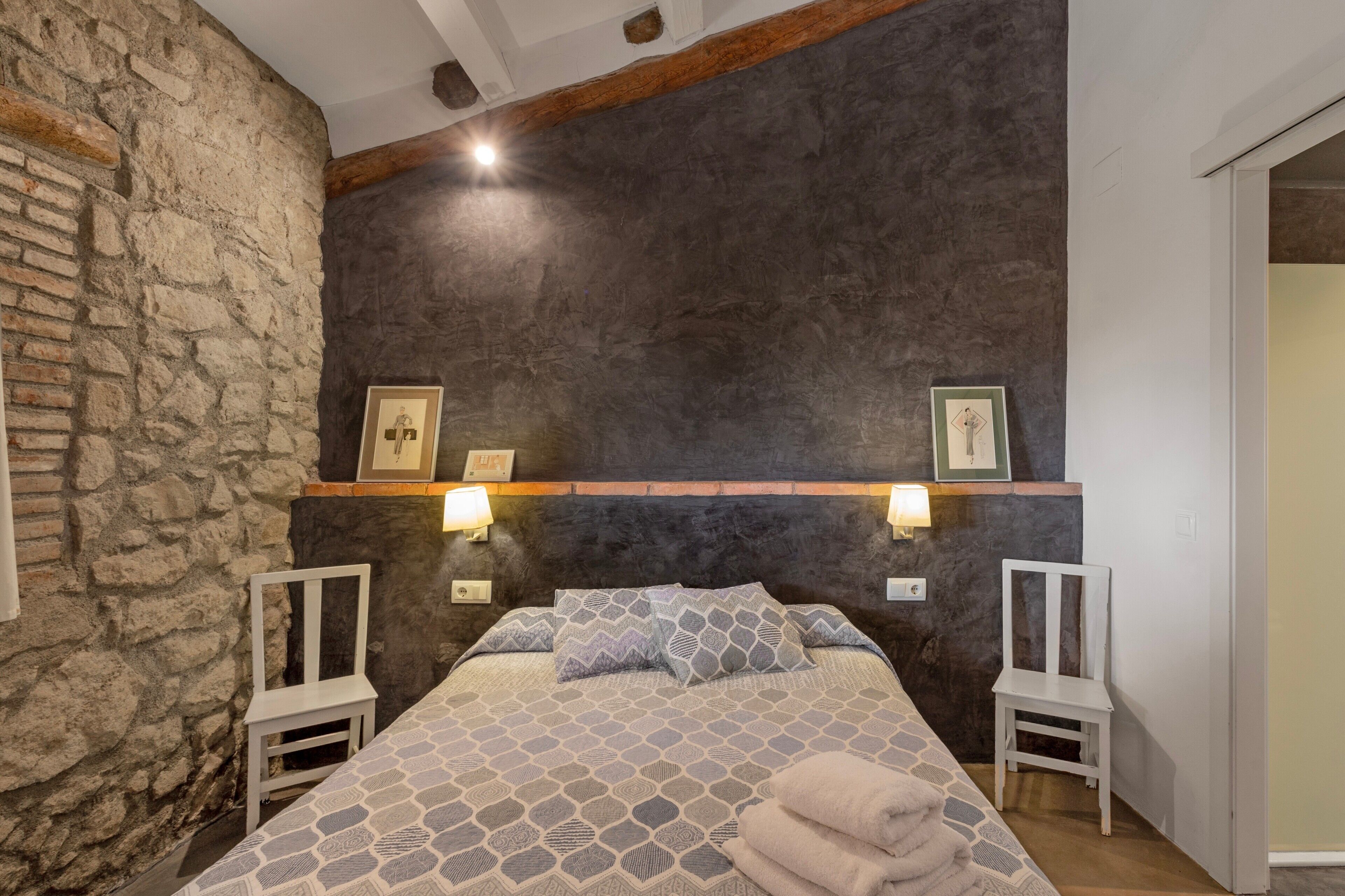 A bedroom with a stone wall, a bed with patterned bedding, two bedside tables with lamps, and a wooden headboard.