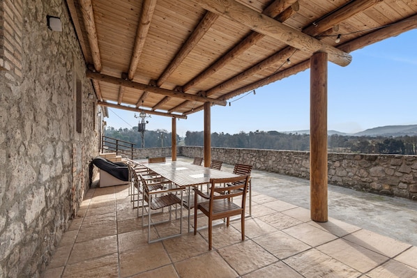 A covered patio with a stone wall, a wooden table and chairs, and a view of the landscape.