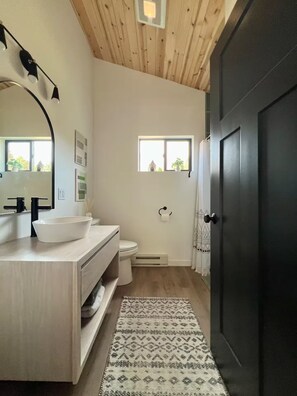 A bathroom with a wooden ceiling, a white sink, a toilet, a small window, and a dark door.