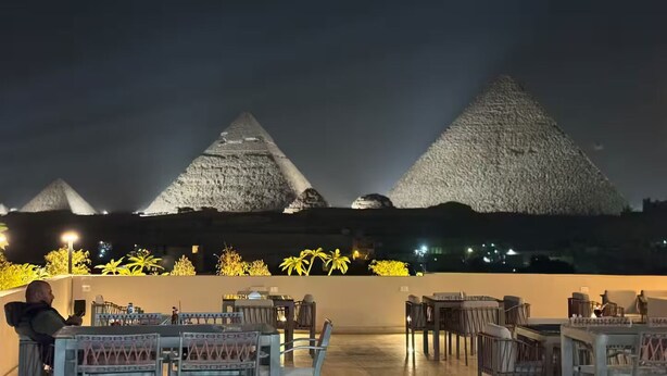 A rooftop terrace with tables and chairs, overlooking three large pyramids. - Sands of Majesty (Al Haram)