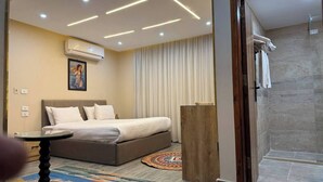 A modern hotel room with a bed, a small table, a shower area, and a painting on the wall.