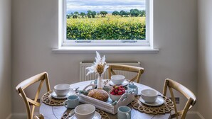 A dining table with a view of a field through a window, surrounded by wooden chairs and a variety of tableware.
