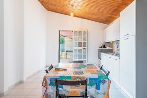 Un espace repas avec une table recouverte d’une nappe à motifs, un plafond en bois et une cuisine dotée de meubles et d’appareils électroménagers blancs.