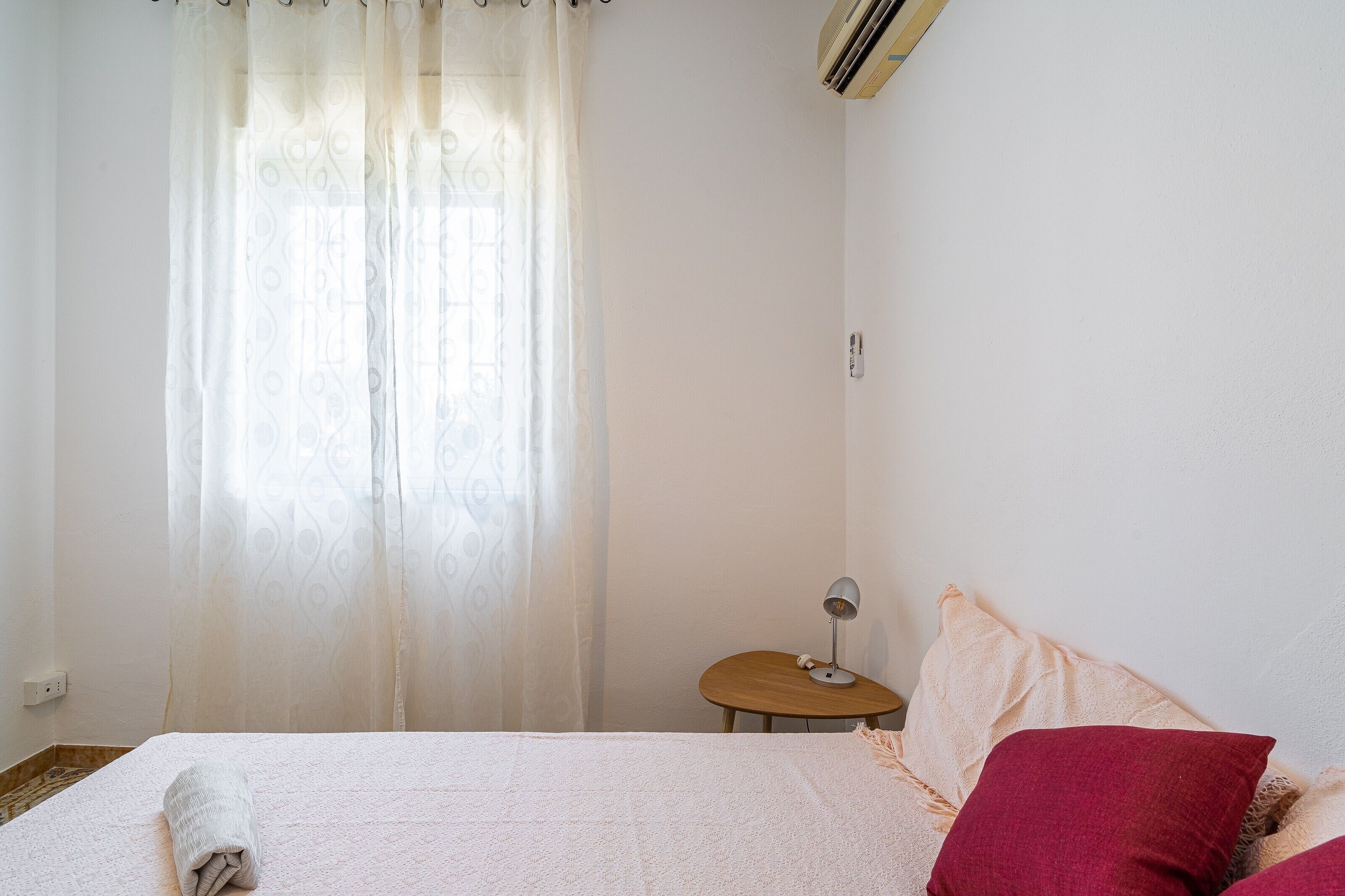 A bedroom with a bed, a bedside table with a lamp, a window with curtains, and a wall-mounted air conditioner.
