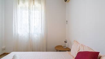 A bedroom with a bed, a bedside table with a lamp, a window with curtains, and a wall-mounted air conditioner.