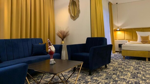 Una habitación de hotel con un sofá azul, dos sillones, una mesita y una cama con cabecero amarillo.