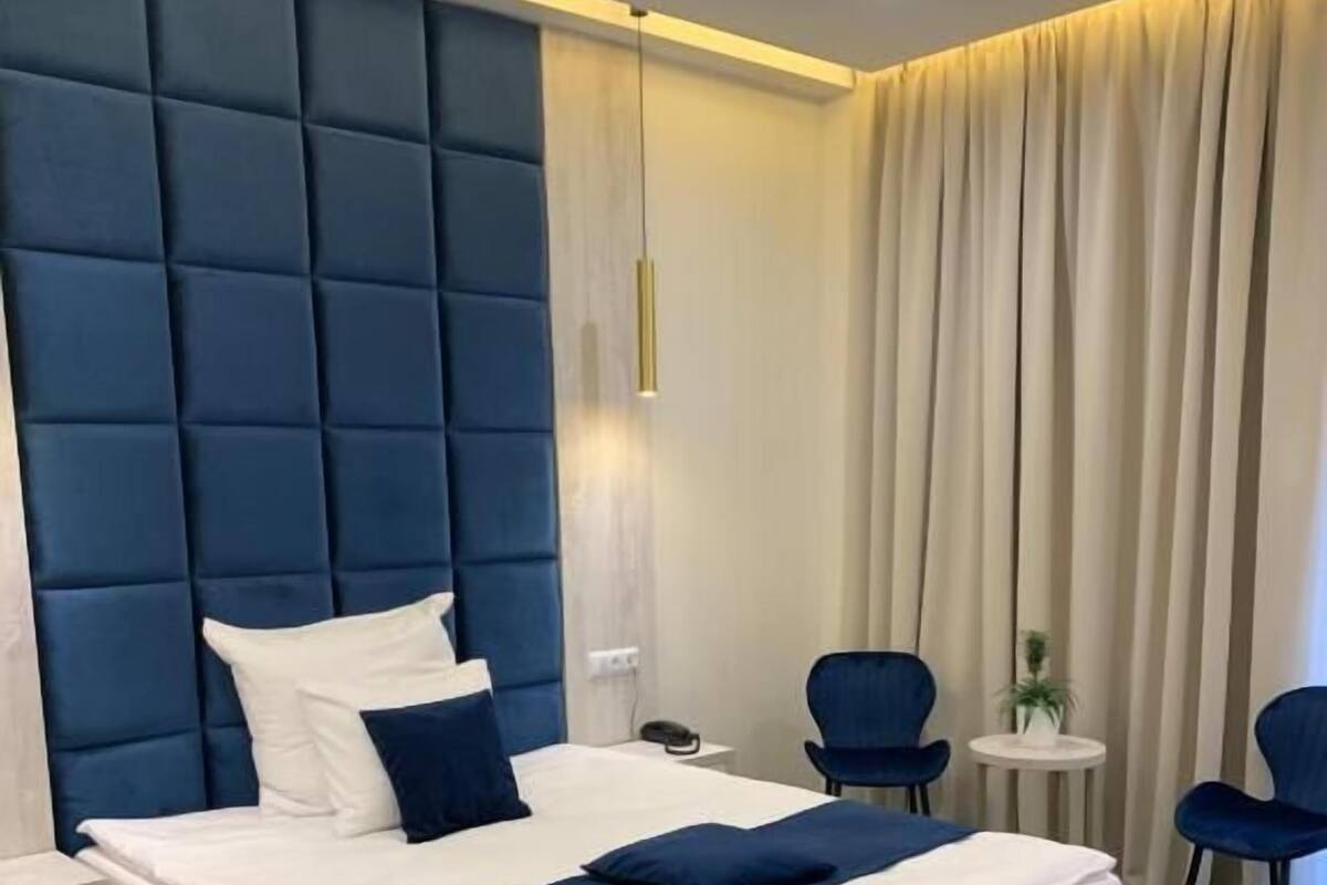 Ein modernes Hotelzimmer mit einem großen Bett, einem Nachttisch, einem kleinen runden Tisch, zwei Stühlen und einem großen Fenster mit Vorhängen.
