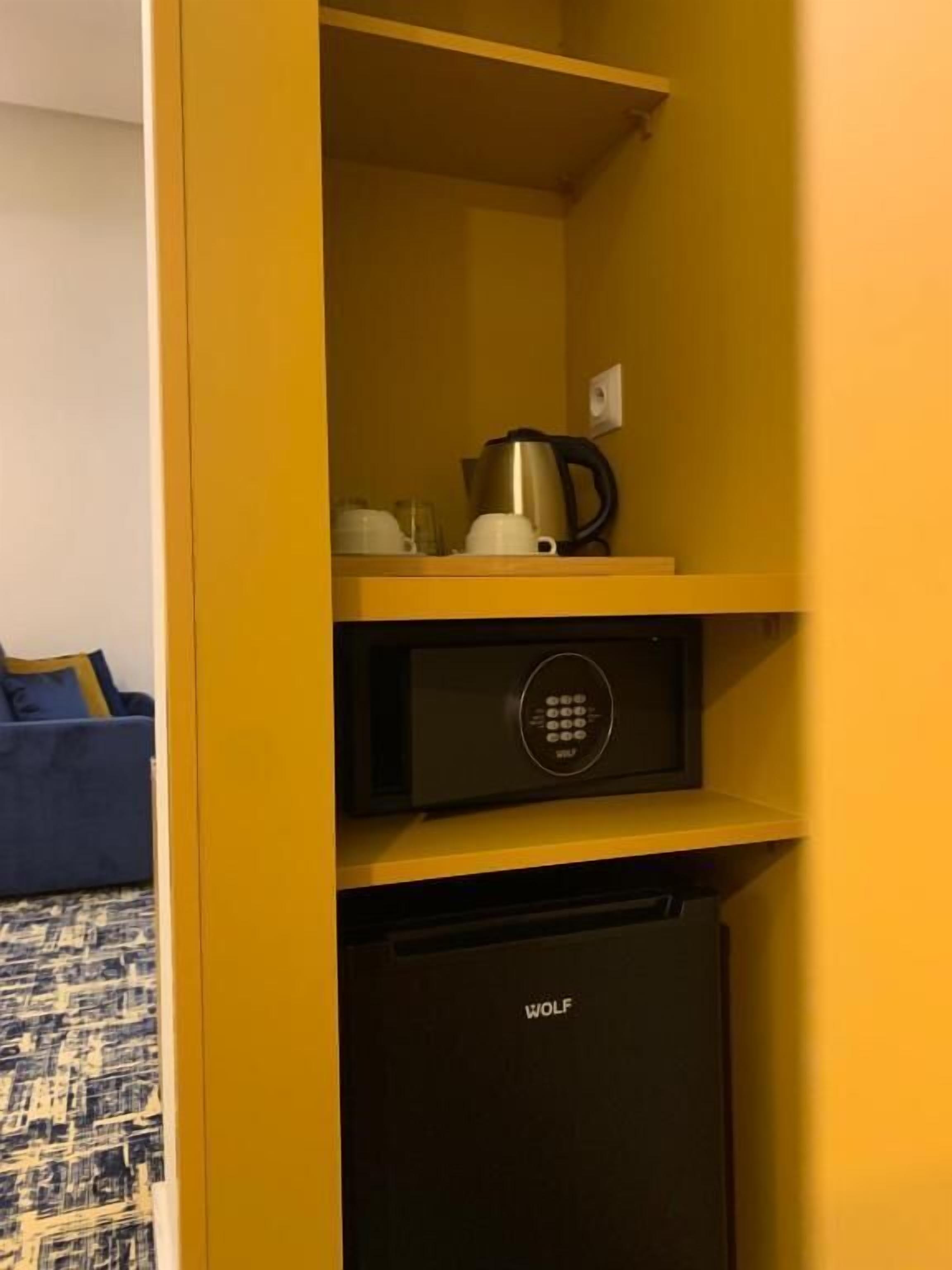 A compact kitchen area with a microwave, kettle, and coffee maker on a yellow shelf, and a black refrigerator below.