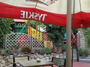 An outdoor seating area with a table and chairs, a red and white canopy, and a colorful playground in the background. - Centrum Piramida (Boleslawiec)