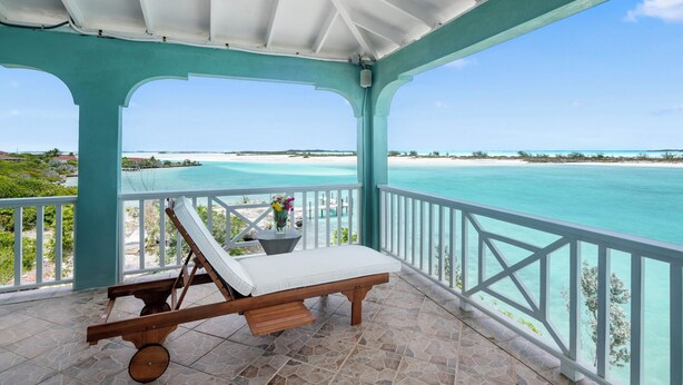 A covered balcony with a view of the ocean, a lounge chair, and a small table with flowers. - Panoramic Ocean Views, Private Beach, Dock, Pickleball, and Housekeeping (Rolletown)