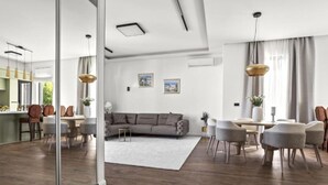 Un salon moderne avec un canapé gris, une table à manger et des chaises. On y trouve une grande fenêtre avec des rideaux, un plafond blanc et un plancher en bois.