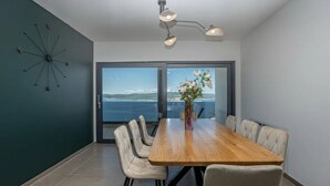 A modern dining room with a wooden table and chairs, a vase of flowers, and a view of the sea through large windows.