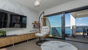 A modern living room with a large TV, a wooden bench, a round marble table, a wicker chair, and a bookshelf.