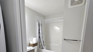 A bathroom with a toilet, a shower with a black and white striped shower curtain, a white sink with a basket of toiletries, and a white toilet paper holder. - Modern 1-Bedroom Garden Suite | Above Ground Suite | Side Parking | Chappelle (Edmonton)