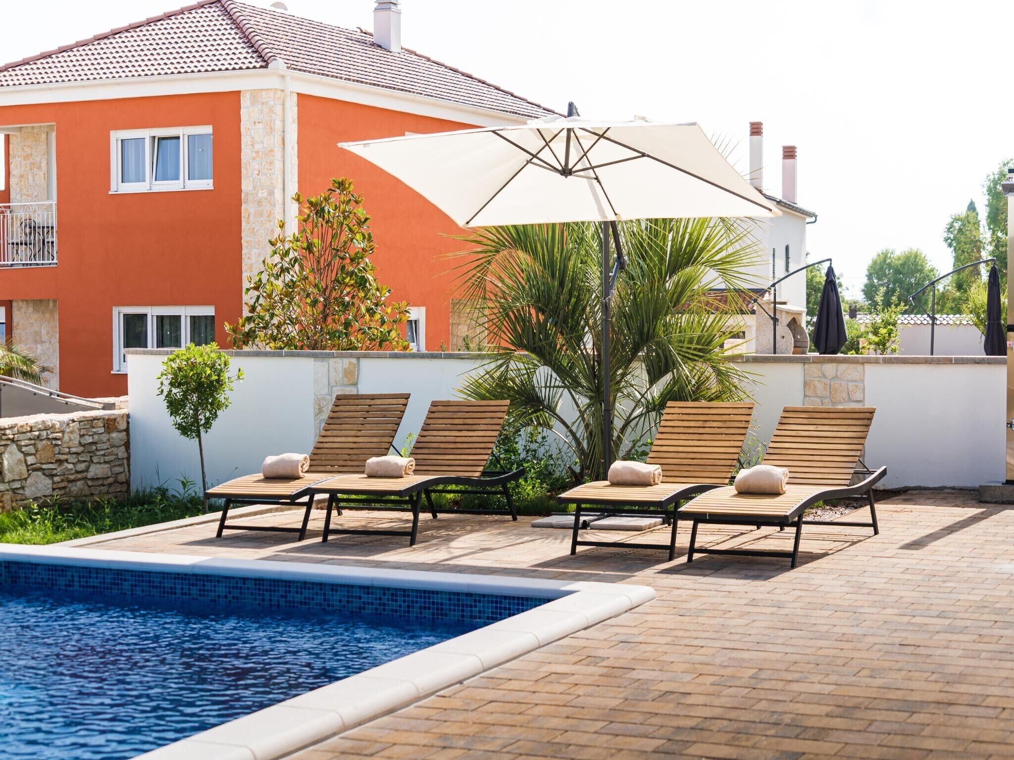Un espace aménagé au bord de la piscine, avec des chaises longues et un parasol, un mur en pierre et un bâtiment doté d’un toit en tuiles.