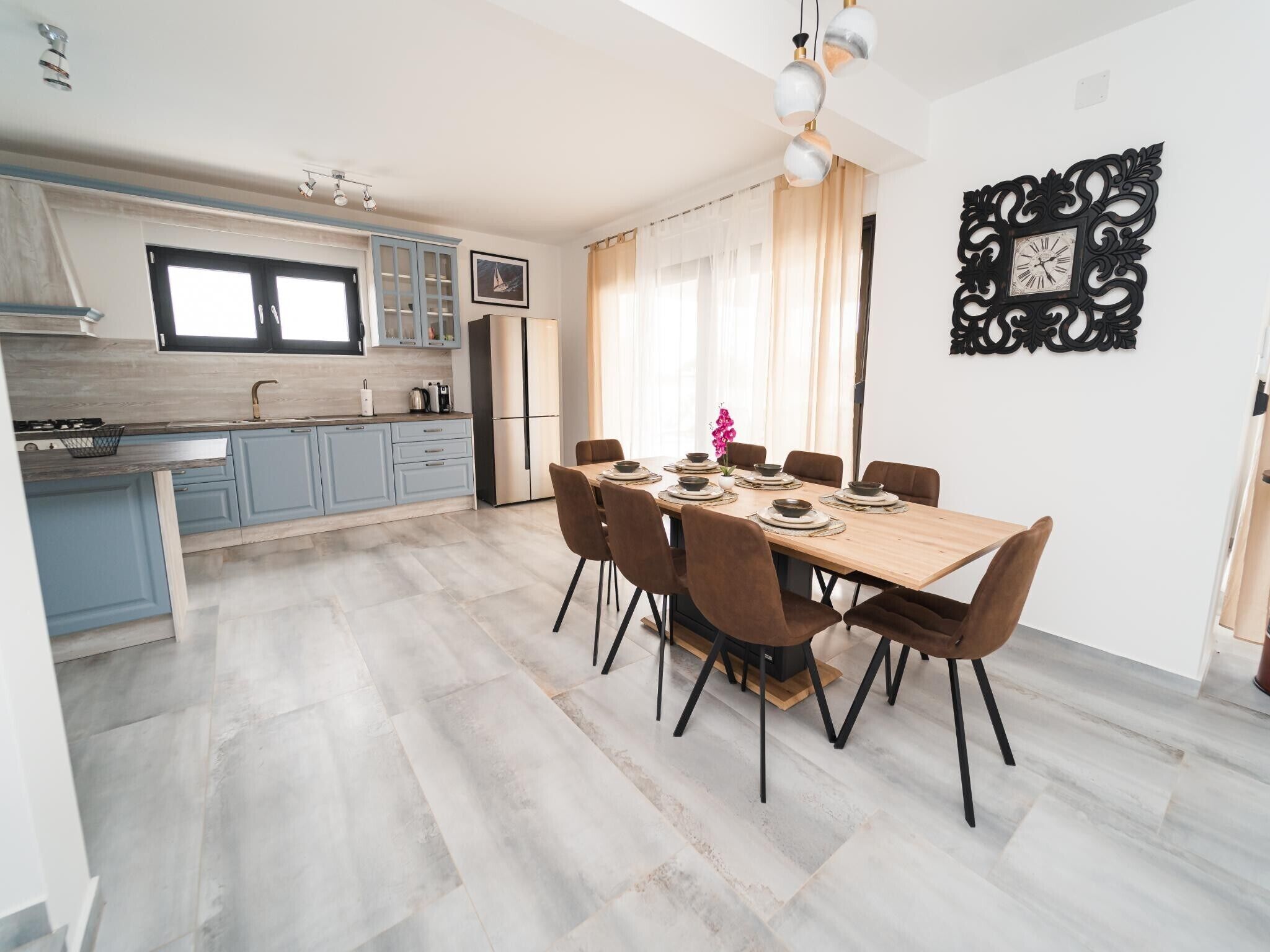 A modern kitchen with light blue cabinets, a central island, and a dining area with a wooden table and brown chairs.