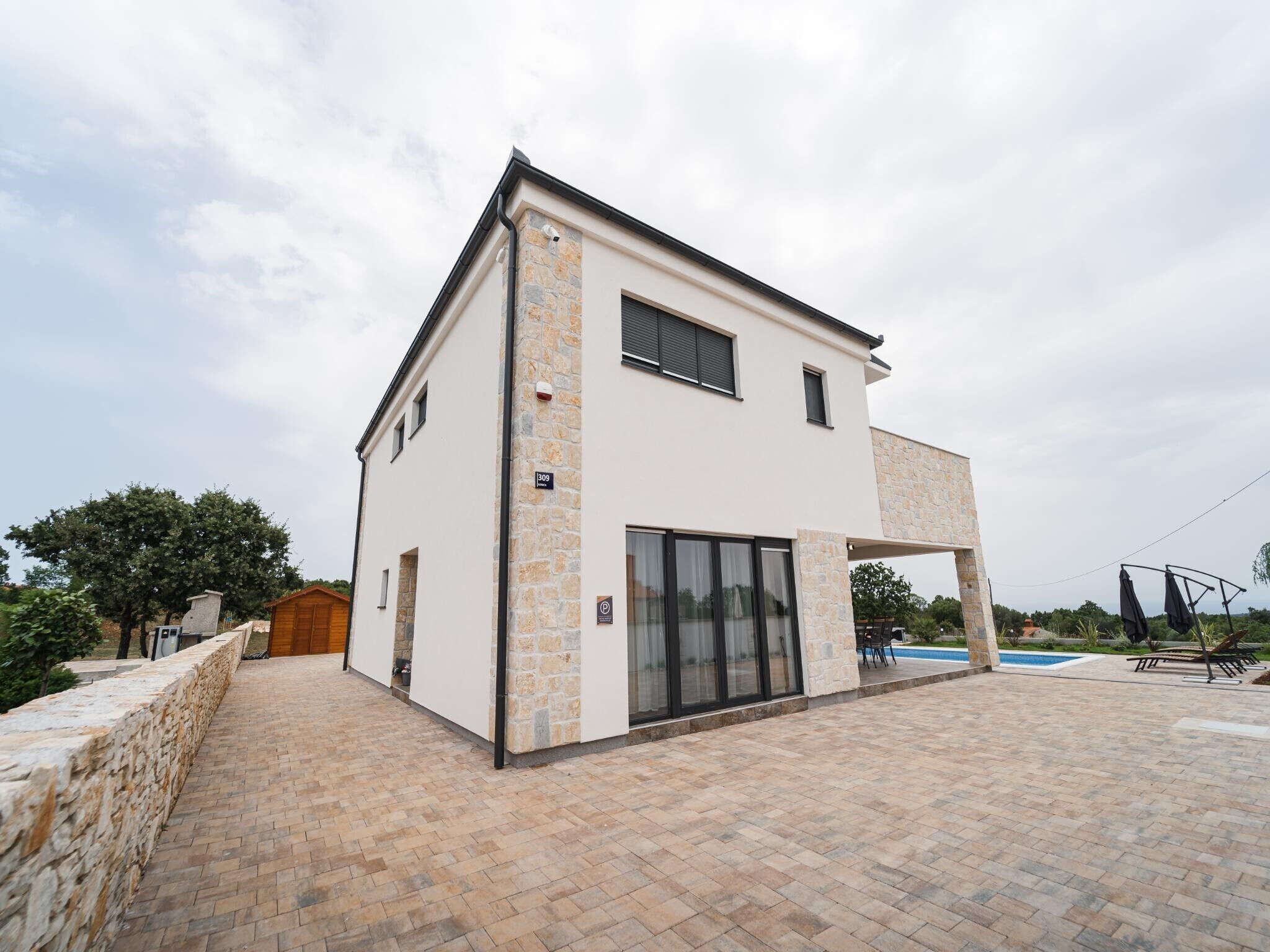 Une maison moderne à deux étages, dotée d’une façade en pierre, d’une grande porte d’entrée en verre et d’un espace piscine visible au fond.