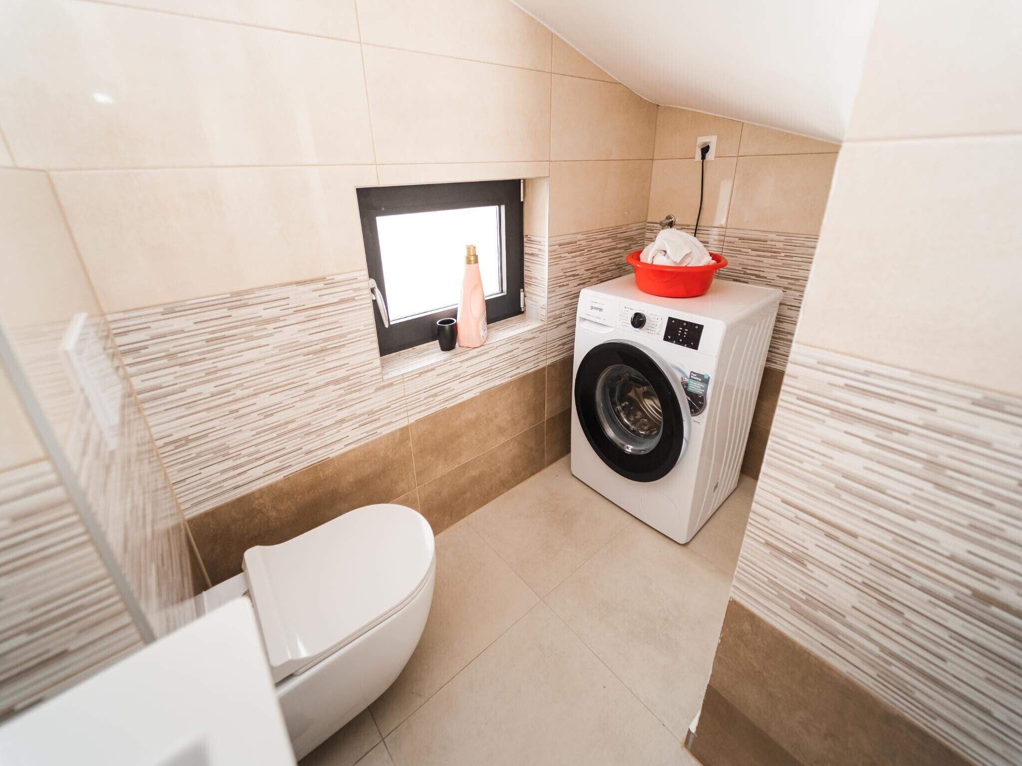 A compact bathroom with a washing machine, a toilet, and a small window.