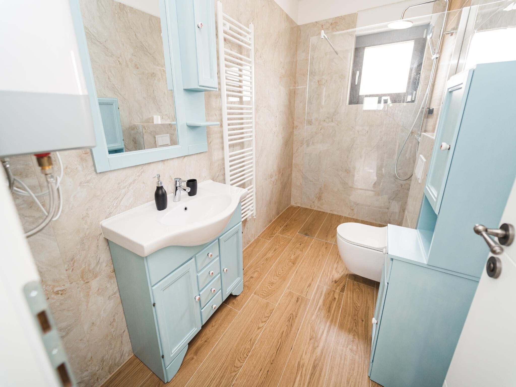 Een moderne badkamer met een houten vloer, een wit wastafelmeubel met zwarte zeepflessen, een wit toilet en een blauwe kast.