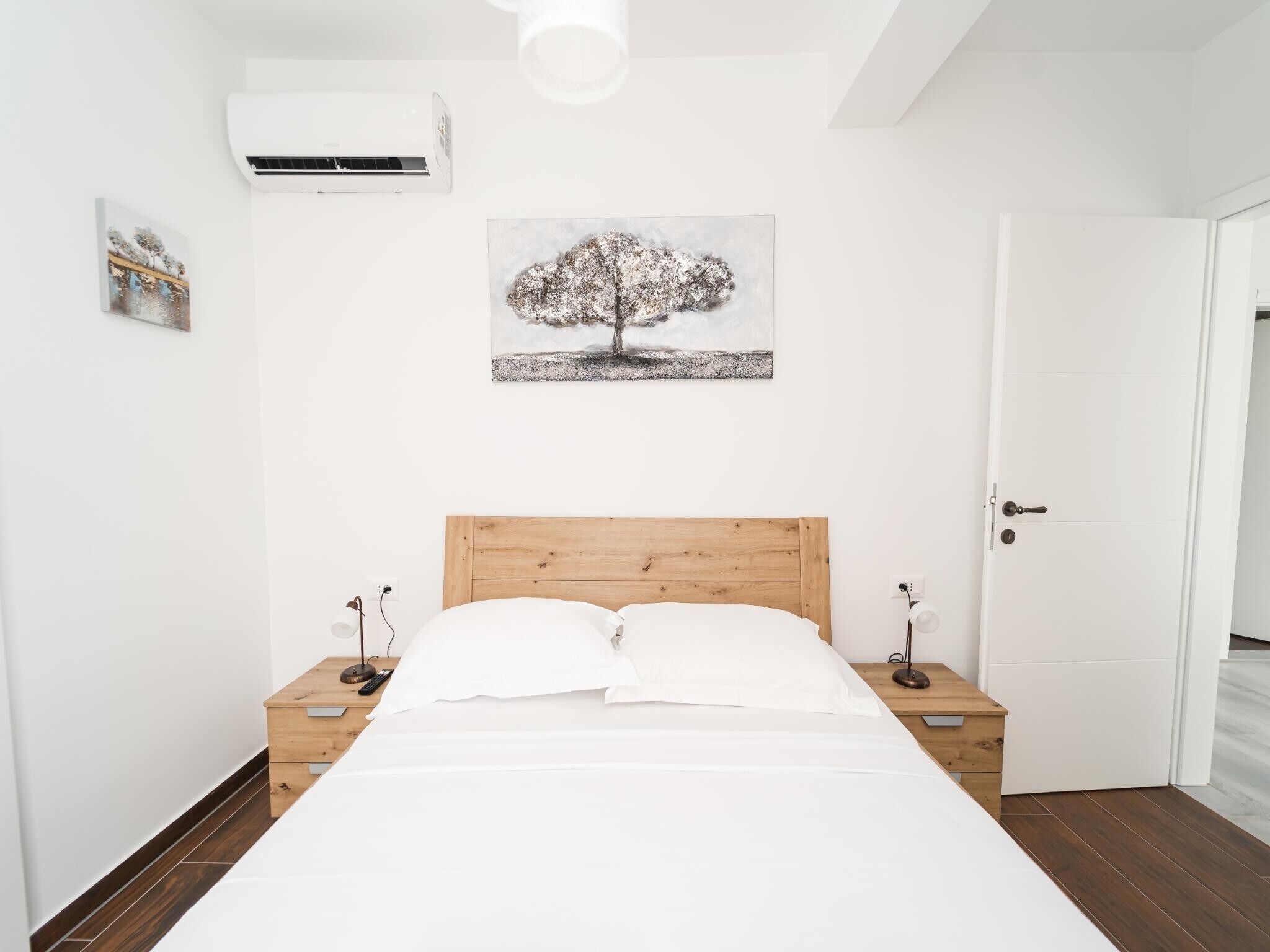 A bedroom with a wooden headboard bed, white bedding, bedside tables with lamps, a wall-mounted air conditioner, and a framed picture of a tree.