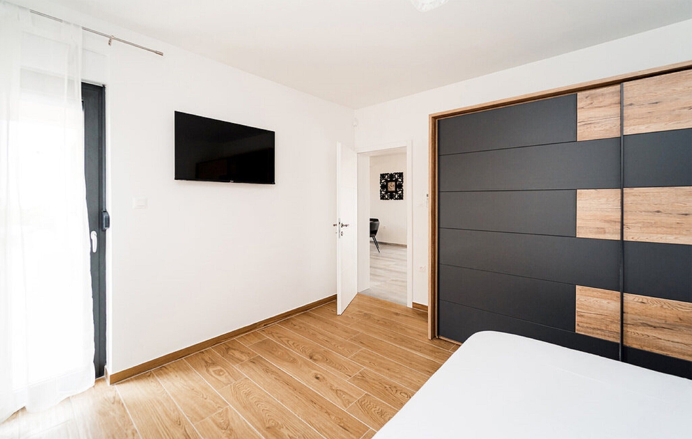 A modern bedroom with a wooden floor, a large wardrobe with a black and wood panel design, and a flat-screen TV mounted on the wall.