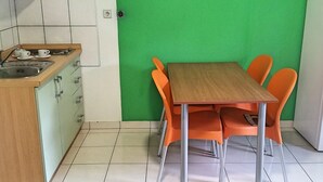 Una pequeña cocina con lavamanos, armarios y pared verde. Hay una mesa de madera con cuatro sillas naranjas.