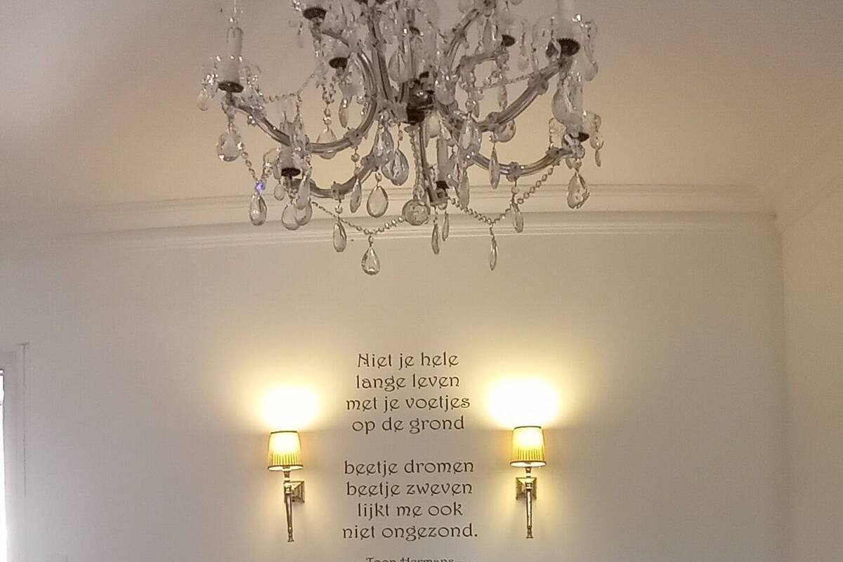 A bedroom with a chandelier, two wall-mounted lamps, a bed with a green blanket, and a quote on the wall.