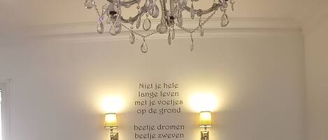 A bedroom with a chandelier, two wall-mounted lamps, a bed with a green blanket, and a quote on the wall.