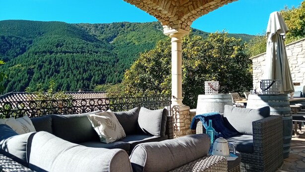 A terrace with a sofa, a stone pillar, and a view of a forested mountain. - Villa Montagnes (Valgorge)
