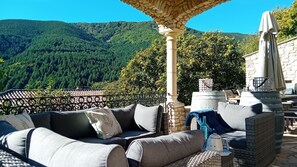 A terrace with a sofa, a stone pillar, and a view of a forested mountain. - Villa Montagnes (Valgorge)