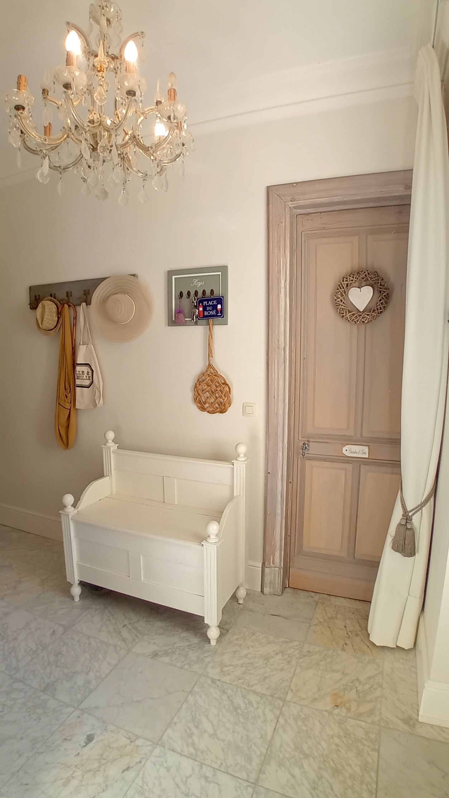 Quarto com cama branca, um candelabro, porta com decoração em formato de coração e um letreiro fixado na parede.