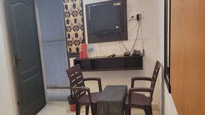 A small, compact room with a sofa, two chairs, a table, a TV mounted on the wall, and a shelf with a few items. - HOTEL I PLAZA (Madurai)