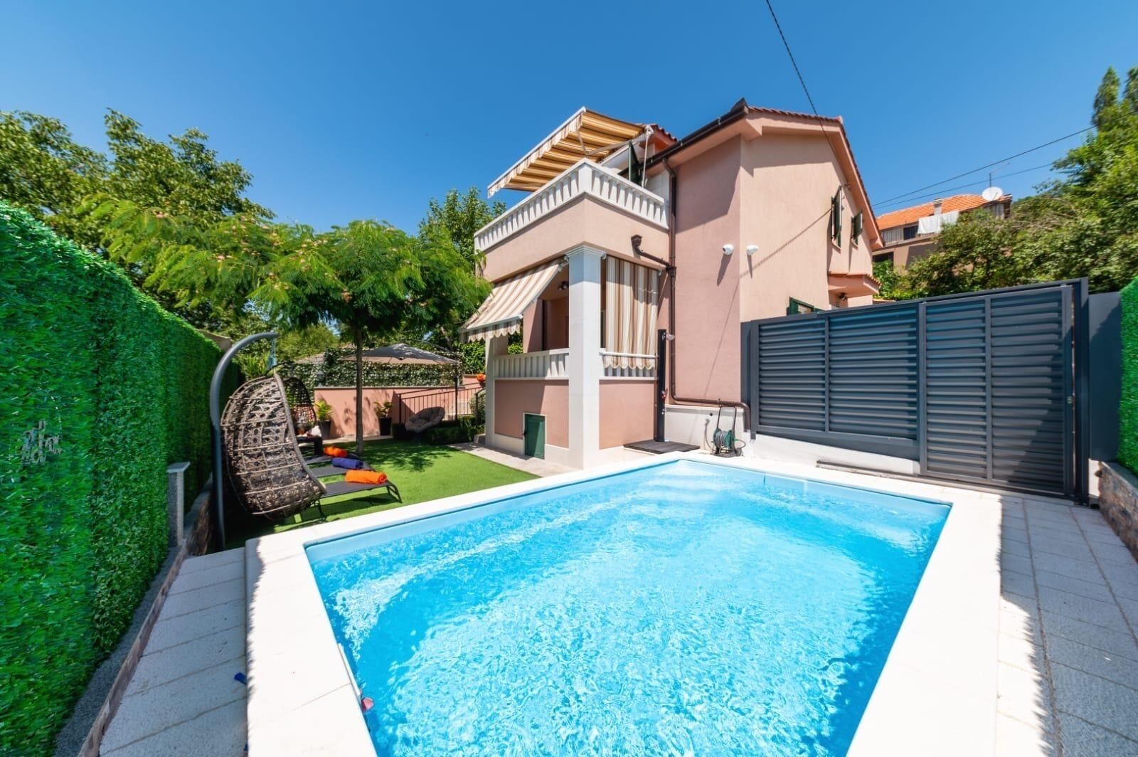A swimming pool with a tiled deck, a two-story house with a balcony, and a green hedge.