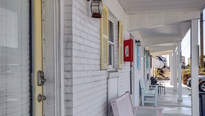 A covered porch with a white brick wall, yellow shutters, and a red fire extinguisher. There are two light-colored benches and a white boat in the background. - Walk to Shore: Atlantic Beach Condo w/ Pool Access (Atlantic Beach)