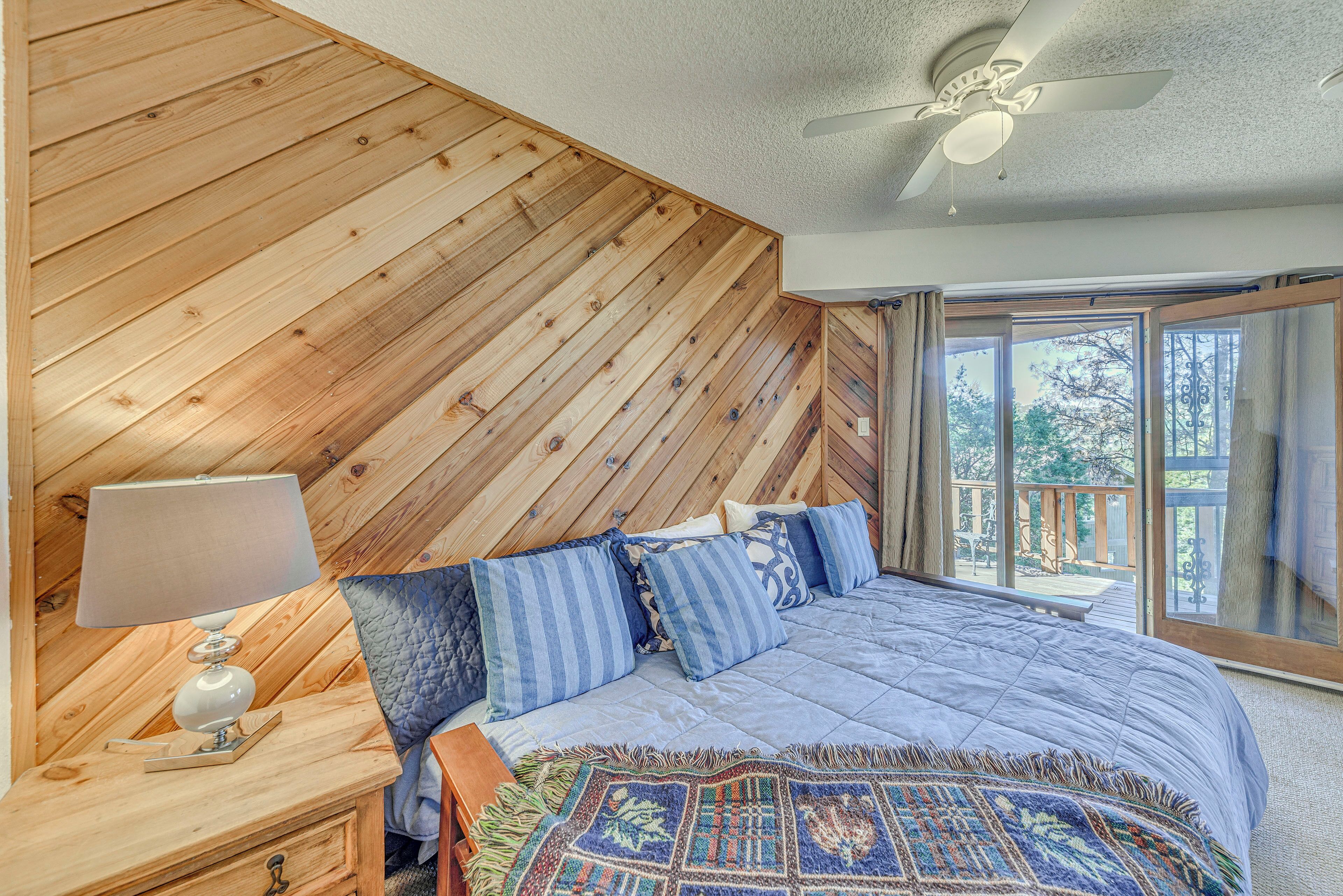 A bedroom with a wooden sloped ceiling, a large bed with blue bedding, a nightstand with a lamp, and a view of the outdoors through a large window.